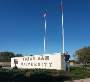 Stypendysta opierający się o tablicę z nazwą "Texas A&M University". W tle widać dwie flagi.